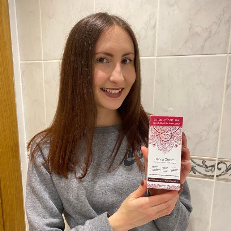 Smiling woman holding up Tint of Nature Henna Cream mahogany red semi-permanent hair dye after using the colour